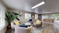 Located at 700 SW Eastman Pkwy in Gresham, Oregon, this single-floor community space offers a well-designed, open layout that seamlessly connects a lounge area, dining space, and kitchenette. The lounge is furnished with contemporary, mid-century-inspired seating — including plush upholstered armchairs and a distinctive glass-top coffee table with a natural wood base — arranged atop a patterned area rug. Multiple windows fitted with white horizontal blinds fill the room with abundant natural light and provide views of the surrounding neighborhood. Tasteful framed artwork and a round wood-framed mirror add character to the neutral-toned walls, while large indoor plants bring a touch of warmth and life to the space. Durable wood-look flooring runs throughout, providing a cohesive and easy-to-maintain surface. The dining area features a solid wood table with matching chairs and benefits from a large sliding glass door with vertical blinds, offering direct access to an outdoor patio area and views of lush greenery. A skylight above further enhances the brightness of this corner. The kitchenette is clean and functional, outfitted with white shaker-style cabinetry, grey countertops, a stainless steel refrigerator, microwave, and a sink — making the space well-equipped for gatherings and events.