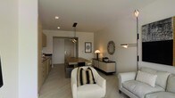 This single-floor space presents an open-plan design that unifies the kitchen, dining, and living areas into one cohesive environment. The kitchen, positioned along one wall, features light wood cabinetry and integrated appliances, offering a functional yet visually streamlined aesthetic. A rectangular dining table with a bench seat and matching chairs sits at the heart of the space, illuminated by a sleek pendant light fixture suspended from the ceiling. Recessed lighting throughout contributes to an evenly lit, contemporary atmosphere.

The living area is anchored by a pair of plush white armchairs, one adorned with a striped accent cushion, alongside a light-toned sofa that reinforces the space's neutral color palette. A low-profile sideboard lines one wall, styled with decorative objects and topped by a warm-toned table lamp. An ornate round mirror with an intricate frame adds a subtle decorative contrast above the sideboard. A framed artwork nearby completes the refined, understated aesthetic. Light-toned flooring runs throughout, tying the open layout together with a sense of continuity and understated elegance.