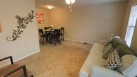Located at 1815 Rosemont Dr in Greenville, NC, this single-floor residence offers a welcoming and functional layout designed for everyday comfortable living. The open living and dining area serves as the heart of the home, where soft, neutral-toned walls create a calm and inviting ambiance. Wall-to-wall carpeting in a warm beige adds a cozy underfoot feel and ties the space together cohesively. The dining area is thoughtfully positioned adjacent to the living space, allowing for easy flow between the two zones — ideal for both casual meals and entertaining. Ceiling lighting fixtures illuminate both areas effectively, with a decorative pendant light adding a subtle design touch above the dining space. Clean white baseboards provide a crisp contrast against the neutral walls, contributing to a tidy and well-maintained aesthetic. The layout is straightforward and practical, making the most of the available floor space. Overall, this residence presents a simple yet comfortable living environment with a consistent, neutral design palette that offers a great foundation for personalization.