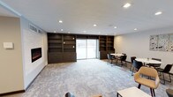 This single-floor space is designed as a contemporary communal lounge or gathering area, blending functional design with a clean, modern aesthetic. The room is anchored by a striking floor-to-ceiling dark wood built-in shelving unit that spans the length of one wall, offering generous storage and display space through a combination of open shelves and closed lower cabinets. At its center, a sliding glass door floods the interior with natural light and provides a direct connection to the outdoors. An electric fireplace is integrated into the adjacent wall, adding a warm, inviting focal point to the space. The flooring features a patterned carpet in soft neutral tones with subtle gold accents, contributing texture and visual interest underfoot. Recessed lighting is distributed evenly across the white ceiling, ensuring bright, uniform illumination throughout. A casual dining or meeting area is situated to one side of the room, furnished with a round white table and contemporary dark chairs, making it well-suited for small group gatherings or collaborative use. The overall ambiance is professional yet welcoming, with a neutral color palette of whites and dark wood tones creating a refined, versatile environment appropriate for a variety of uses.
