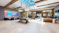 Located at 112 Bilby Road in Hackettstown, NJ, this expansive single-floor amenity space offers a generously sized, open-concept layout designed for both recreation and relaxation. The architectural design blends rustic and contemporary elements seamlessly, with exposed wooden ceiling beams and large skylights that flood the interior with abundant natural light, creating a bright and welcoming atmosphere throughout.

The main gathering area serves as the centerpiece of the space, featuring a full-size billiards table, a foosball table, and a large-format display screen, all set against light-toned hardwood flooring. Sculptural pendant lighting adds a distinctive design flair overhead. Adjacent to the game area, a stylish lounge zone includes a tiled fireplace wall flanked by built-in shelving, comfortable modern seating, and curated décor.

A dedicated fitness center is enclosed behind sleek black-framed glass walls, providing a well-equipped workout area with ample equipment and mirror-lined walls that enhance the sense of space. Nearby, a sophisticated bar area with upholstered counter seating and a warm gold-toned bar front opens directly to an outdoor courtyard through full-height glass doors. The reception area features a textured tile accent wall, bold artwork, and a stylish welcome desk. A smart parcel locker system and an ornate metal screen partition round out this thoughtfully designed, multifunctional facility.