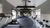 This well-appointed fitness center is housed within a single-story structure distinguished by its striking vaulted ceilings adorned with dark, rustic wood beams that add architectural character to the space. Two ceiling fans are suspended from the peak of the vault, ensuring steady air circulation throughout the room during workouts.

The interior is finished in a cool grey palette, creating a focused and energizing atmosphere. Large windows line both sides of the room, flooding the space with natural light and offering views of the surrounding greenery, while maintaining a sense of openness. A wall-mounted television is positioned at the far end of the room, visible from most workout areas.

The flooring consists of durable dark rubber matting, purpose-built for fitness use. The equipment layout is thoughtfully organized, with cardio machines—including ellipticals and stationary bikes—arranged along one side, while a comprehensive selection of white cable and weight machines occupies the opposite side. The generous floor plan provides ample space between equipment, allowing for comfortable movement and a variety of workout routines without feeling crowded.