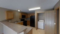 This single-floor residence is designed with practicality and comfort in mind. The kitchen serves as a central hub of the home, outfitted with warm honey-toned wood cabinetry that provides generous storage space. Black appliances, including a refrigerator, electric range, and over-the-range microwave, offer a cohesive and modern look. A double-basin sink is conveniently positioned along the counter, complemented by a light-toned countertop that extends along the perimeter, providing ample workspace. A breakfast bar-style peninsula adds additional counter space and creates a natural divide between the kitchen and the adjacent living area. The light tile flooring throughout the kitchen lends a clean, low-maintenance finish to the space. A notable feature of this residence is the dedicated in-unit laundry closet, located just off the kitchen. Equipped with a washer and overhead storage rod, this space adds significant day-to-day convenience. Overhead lighting fixtures illuminate the kitchen efficiently. The overall ambiance of the space is warm and functional, with a neutral color palette that provides a versatile backdrop suitable for a variety of décor styles.