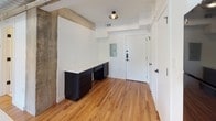 This 1-bedroom, 1-bathroom unit showcases a compelling mix of industrial heritage and contemporary design. Warm hardwood floors run throughout the space, complementing the exposed concrete columns and beams that lend authentic character to the interior. The open-concept kitchen serves as a centerpiece, featuring bold matte black cabinetry stacked to the ceiling, black appliances including a refrigerator, electric range, and microwave, and a generous white quartz island with an undermount sink. Pendant lights hang above the island, adding a polished touch, while exposed ductwork overhead reinforces the loft-inspired aesthetic. The bedroom area includes a built-in workspace with dark cabinetry and a white countertop surface, as well as access to a well-organized closet equipped with dual clothing rods and shelving for ample storage. The bathroom continues the sophisticated black-and-white palette with dark cabinetry, white countertops, white tile, and a standout combination tub and shower fitted with striking gold fixtures that add a refined contrast to the otherwise monochromatic scheme. The unit's cohesive design language — raw industrial elements balanced with clean, modern finishes — creates an inviting and distinctive living environment.
