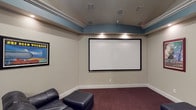 This single-floor home theater space is thoughtfully designed to deliver a true cinematic experience. The centerpiece of the room is a large, wall-mounted projection screen framed in black, positioned for optimal viewing from any seat. Plush black leather seating, including a reclining chair with an ottoman, is arranged to face the screen, creating a comfortable and immersive viewing environment. The carpeted floor in a rich, dark burgundy tone adds warmth and helps with acoustic dampening throughout the space. A distinctive architectural detail is the tray ceiling, accented with a soft teal-blue band that runs along its perimeter, adding a touch of elegance and visual depth. Recessed and in-ceiling lighting illuminate the space evenly, while multiple built-in ceiling speakers suggest a full surround sound audio system. Complementing the theater area is a compact kitchenette equipped with light wood cabinetry, a dark countertop, a sink, and a microwave, making it convenient for entertaining. A door leads to an adjacent area, rounding out this versatile and well-appointed entertainment space.