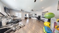 This fitness center offers a thoughtfully designed, open-plan workout space finished with warm hardwood-style flooring and crisp white walls that create a clean, energizing atmosphere. The room is illuminated by recessed lighting and benefited by multiple windows with blinds that allow natural light to filter throughout the space, keeping it bright and inviting.

Dual ceiling fans are mounted overhead, ensuring consistent airflow during workouts. The layout is generous enough to accommodate a wide variety of equipment without feeling crowded, providing ample room for free movement between stations.

The strength training area includes a multi-function cable machine, a weight bench with barbell, and a full dumbbell rack, complemented by three tall framed mirrors along one wall that enhance the sense of space and allow users to monitor their form. A motivational "Harder. Better. Faster. Stronger." wall decal adds an energizing focal point.

The cardio zone features treadmills, an elliptical, a stationary bike, and a rowing machine. Additional accessories include stability balls, medicine balls, resistance tools, and yoga mats. A wall-mounted flat-screen TV and a small storage unit round out the amenities, making this a fully functional and well-organized fitness facility.
