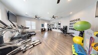 This fitness center offers a thoughtfully designed, open-plan workout space finished with warm hardwood-style flooring and crisp white walls that create a clean, energizing atmosphere. The room is illuminated by recessed lighting and benefited by multiple windows with blinds that allow natural light to filter throughout the space, keeping it bright and inviting.

Dual ceiling fans are mounted overhead, ensuring consistent airflow during workouts. The layout is generous enough to accommodate a wide variety of equipment without feeling crowded, providing ample room for free movement between stations.

The strength training area includes a multi-function cable machine, a weight bench with barbell, and a full dumbbell rack, complemented by three tall framed mirrors along one wall that enhance the sense of space and allow users to monitor their form. A motivational "Harder. Better. Faster. Stronger." wall decal adds an energizing focal point.

The cardio zone features treadmills, an elliptical, a stationary bike, and a rowing machine. Additional accessories include stability balls, medicine balls, resistance tools, and yoga mats. A wall-mounted flat-screen TV and a small storage unit round out the amenities, making this a fully functional and well-organized fitness facility.