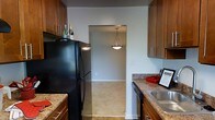 This single-story space includes a well-appointed kitchen designed for everyday functionality and comfort. The kitchen features warm-toned wood cabinetry that extends from floor to ceiling, providing ample storage on both sides of the workspace. Granite-style countertops offer generous prep surfaces, complemented by a stainless steel sink and black appliances, including a full-sized refrigerator and dishwasher. The layout is galley-style, creating an efficient corridor that leads directly into an adjacent room — likely a dining or living area — visible through an open arched passageway. That neighboring space is illuminated by a pendant light fixture with a classic cone shade, suspended from the ceiling, adding a subtle decorative touch. The light-toned flooring flows seamlessly between the two areas, contributing to a cohesive and open feel. Neutral gray walls throughout maintain a clean, understated backdrop that allows the wood cabinetry and warm countertop tones to stand out. Overall, the space presents a practical yet inviting interior with a straightforward, traditional design aesthetic.