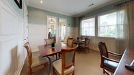 Located at 501 Burke Dr in Hinesville, Georgia, this single-floor space exudes a polished, welcoming character through its thoughtfully curated interiors. The design palette features soft sage-teal upper walls paired with crisp white wainscoting panels, creating a refined and cohesive aesthetic throughout. Crown molding adds an additional touch of classic elegance to each room.

The office area is well-appointed with a substantial dark cherry executive desk, a swivel chair, and a literature rack stocked with informational materials. Framed nautical-themed artwork adorns the walls, and a table lamp provides warm ambient lighting. A meeting table with upholstered chairs offers a comfortable setting for consultations or small group gatherings.

The dining area features multiple dark cherry wood tables, each accompanied by upholstered side chairs in neutral tones. Large windows dressed with draping curtains and horizontal blinds allow generous natural light to fill the room, while offering views of the surrounding palm-lined landscape. The open doorways between areas promote a seamless flow throughout the space.

Neutral carpet flooring, recessed lighting, and carefully placed greenery complete the inviting atmosphere, making this a functional and aesthetically pleasing environment suited for both professional and social interactions.