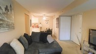 This single-floor residence offers a welcoming and efficient layout that seamlessly connects the main living areas. The living room serves as the heart of the space, featuring plush carpeting, warm neutral-toned walls, and a ceiling fan with a light fixture that provides both comfort and illumination. A large sectional sofa anchors the room, creating an inviting area for relaxation.

The open-concept design flows naturally from the living room into a casual dining area, which features a compact dining table and chairs — ideal for everyday meals. Just beyond, the kitchen is neatly appointed with white cabinetry, light countertops, and essential appliances, offering a clean and organized cooking space that remains visually connected to the rest of the living area.

A set of large closet doors is prominently featured along one wall, providing generous built-in storage that contributes to the unit's practicality. The consistent warm, beige color palette throughout creates a cohesive and cozy ambiance. The single-floor layout ensures easy accessibility and a straightforward flow between all areas of the space, making it a comfortable and functional place to call home.