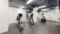 Located at 200 Dufferin Street in Toronto's vibrant King West neighbourhood, this fitness facility offers a dedicated workout space across a single, open floor. The gym features an industrial-style aesthetic with an exposed ceiling revealing overhead pipes and ductwork, painted in a dark grey finish and illuminated by rows of bright fluorescent strip lighting that ensure the entire space is well-lit.

The flooring throughout is durable speckled rubber, ideal for high-traffic fitness use and providing both grip and impact absorption. White-painted concrete block walls frame the perimeter, lending the space a clean, no-frills atmosphere. Structural columns are painted white, helping to visually segment the floor into distinct workout zones.

The cardio section is well-appointed, featuring multiple treadmills, elliptical trainers, and upright and recumbent stationary bikes, catering to a variety of cardiovascular training preferences. A dedicated strength and resistance training area includes a multi-station cable machine, with large wall-mounted mirrors reflecting the equipment and enhancing the sense of depth throughout the space. A decorative wall mural adds a subtle design accent to one section of the gym.

The layout is open and functional, allowing users to move freely between equipment zones. The space is rounded out by a clearly marked exit door, ensuring safe and convenient access.
