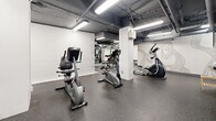 Located at 200 Dufferin Street in Toronto's vibrant King West neighbourhood, this fitness facility offers a dedicated workout space across a single, open floor. The gym features an industrial-style aesthetic with an exposed ceiling revealing overhead pipes and ductwork, painted in a dark grey finish and illuminated by rows of bright fluorescent strip lighting that ensure the entire space is well-lit.

The flooring throughout is durable speckled rubber, ideal for high-traffic fitness use and providing both grip and impact absorption. White-painted concrete block walls frame the perimeter, lending the space a clean, no-frills atmosphere. Structural columns are painted white, helping to visually segment the floor into distinct workout zones.

The cardio section is well-appointed, featuring multiple treadmills, elliptical trainers, and upright and recumbent stationary bikes, catering to a variety of cardiovascular training preferences. A dedicated strength and resistance training area includes a multi-station cable machine, with large wall-mounted mirrors reflecting the equipment and enhancing the sense of depth throughout the space. A decorative wall mural adds a subtle design accent to one section of the gym.

The layout is open and functional, allowing users to move freely between equipment zones. The space is rounded out by a clearly marked exit door, ensuring safe and convenient access.