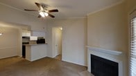This 1-bedroom, 1-bathroom unit is thoughtfully laid out across a single floor, offering a comfortable and functional living environment. The main living area features carpeted flooring, crown molding, and a classic white-mantled fireplace with a dark surround, serving as a natural focal point of the room. A ceiling fan with an integrated light fixture is centrally positioned overhead, providing both air circulation and ambient lighting. The living space flows seamlessly into the kitchen, which is visible through an open pass-through counter. The kitchen is equipped with white cabinetry, a dark countertop, and black appliances, including a microwave. Neutral-toned walls throughout the unit create a clean, versatile backdrop. A doorway from the living area leads to the private bedroom and bathroom, ensuring a clear separation between living and sleeping spaces. The overall layout is efficient and well-organized, making thoughtful use of the available square footage. Crown molding details add a subtle architectural refinement, contributing to the unit's polished and well-maintained appearance.