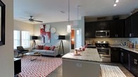 This single-floor residence showcases a thoughtfully designed open-concept layout that seamlessly connects the kitchen and living areas into one cohesive, inviting space. The kitchen is appointed with dark espresso cabinetry, sleek stainless steel appliances — including a range, microwave, and additional cooking surface — and a generous granite-topped island. Stylish pendant lights hang above the island, providing both focused task lighting and an eye-catching design element. The living area is warm and well-appointed, featuring hardwood-style flooring accented by a bold geometric area rug in red and white tones. A ceiling fan ensures year-round comfort overhead. Contemporary floor lamps flank a plush sofa accented with red throw pillows, while vibrant wall art adds a pop of color and personality to the neutral, light grey walls. A doorway visible in the background hints at additional rooms beyond, offering a sense of depth and flow throughout the floor plan. The overall ambiance strikes a balance between modern sophistication and comfortable livability, making it an ideal space for both relaxing and entertaining.