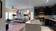 This single-floor residence showcases a thoughtfully designed open-concept layout that seamlessly connects the kitchen and living areas into one cohesive, inviting space. The kitchen is appointed with dark espresso cabinetry, sleek stainless steel appliances — including a range, microwave, and additional cooking surface — and a generous granite-topped island. Stylish pendant lights hang above the island, providing both focused task lighting and an eye-catching design element. The living area is warm and well-appointed, featuring hardwood-style flooring accented by a bold geometric area rug in red and white tones. A ceiling fan ensures year-round comfort overhead. Contemporary floor lamps flank a plush sofa accented with red throw pillows, while vibrant wall art adds a pop of color and personality to the neutral, light grey walls. A doorway visible in the background hints at additional rooms beyond, offering a sense of depth and flow throughout the floor plan. The overall ambiance strikes a balance between modern sophistication and comfortable livability, making it an ideal space for both relaxing and entertaining.