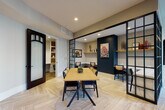 This thoughtfully designed single-floor amenity space blends functionality with sophisticated style across several distinct zones. At its heart is an open lounge area anchored by a natural wood communal table set atop striking herringbone-patterned light wood tile flooring. Sleek black-framed glass partition walls divide the space into semi-private meeting alcoves, each accented with textured wallcoverings, deep navy feature walls, floating shelves, and curated artwork, creating a refined yet approachable atmosphere.

Adjacent to the main lounge, a dedicated work nook offers a built-in marble-topped desk, shiplap-paneled walls, integrated open shelving, and warm brass sconce lighting — providing a focused and elegant environment for individual productivity. Plush leather and cane accent chairs are thoughtfully placed throughout, offering flexible seating options.

Teal-painted built-in bookshelves frame a passageway leading into a recreation room equipped with a pool table and pendant lighting, while cozy reading corners with velvet lounge chairs and framed artwork invite quiet relaxation. The space is finished with recessed lighting throughout, contributing to a warm and welcoming ambiance suited for both collaboration and focused work.