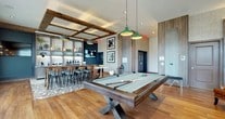 This thoughtfully designed single-floor amenity space masterfully combines industrial aesthetics with contemporary comfort. Exposed brick walls, reclaimed wood ceiling beams, and large steel-framed windows define the architectural character throughout, flooding the interior with abundant natural light and offering views of the surrounding landscape.

The main gathering area centers around a full-size billiards table set atop warm hardwood flooring, complemented by a stylish bar with counter seating on patterned tile. Industrial-style pendant lighting hangs above both zones, reinforcing the space's urban, loft-inspired character. French doors connect the interior to an outdoor patio, enhancing the sense of openness.

A dedicated lounge area provides a more relaxed atmosphere, anchored by a sleek linear fireplace set against a dramatic brick wall with a wall-mounted television above. Plush teal velvet armchairs, a rustic coffee table on casters, and a sculptural chandelier complete this inviting setting.

Tucked within the lounge is an intimate reading nook featuring floor-to-ceiling wooden shelving filled with curated décor and books, paired with rich blue accent chairs on a textured area rug. Together, these distinct zones create a versatile, upscale communal environment ideal for socializing, relaxing, or recreation.