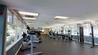 This fitness center is designed to support a comprehensive cardio workout experience within a generously sized, open floor plan. The space is outfitted with rows of modern cardio equipment, including treadmills, elliptical trainers, and stair climbers, all arranged to allow comfortable movement between machines. The rubber flooring provides a durable, cushioned surface suited for high-traffic fitness use.

Large windows line one side of the room, flooding the space with natural light and offering views of the surrounding greenery, creating an open and inviting atmosphere. The drop ceiling with recessed panel lighting ensures consistent, even illumination throughout. Mounted televisions are positioned for visibility from multiple machines, adding convenience for users during longer workouts.

Mirrored walls along one section of the room visually expand the space while allowing users to monitor their form. The overall layout is clean and functional, with a neutral color palette that contributes to a focused workout environment. The facility appears well-maintained, making it a practical and accessible option for cardiovascular fitness.