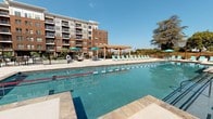 Located at 1000 Goddin St in Richmond, Virginia, this single-story amenity facility is part of a contemporary multi-story residential community characterized by its classic brick exterior and modern design. The space is thoughtfully designed to support an active lifestyle, offering residents access to both indoor fitness facilities and an expansive outdoor recreation area.

The fitness center is well-equipped with a comprehensive selection of cardio machines, including treadmills, ellipticals, stationary bikes, and incline trainers arranged across a clean, open floor plan. Recessed lighting and ceiling fans maintain a comfortable environment, while large windows with blinds allow natural light to fill the room. The interior finishes are crisp and modern, featuring white walls and clean-lined baseboards throughout.

The space also provides direct access to the outdoor pool area, visible through a glass-paneled door. The resort-style pool is generously sized with lap lanes, surrounded by a spacious sun deck lined with lounge chairs and shaded by a wooden pergola with teal umbrellas. The pool area is framed by mature trees and overlooks the broader residential complex, creating an inviting outdoor retreat well-suited to Richmond's warm seasons.