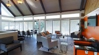 This single-floor communal space makes a striking first impression with its dramatic vaulted ceiling adorned with rich, dark-stained exposed wood beams, lending the interior a warm yet architecturally refined character. The ceiling's angular pitch creates a sense of generous vertical space throughout the room.

Floor-to-ceiling windows and transom glazing line the perimeter walls, flooding the interior with natural light and offering lush, tree-lined views of the surrounding outdoor area. Sheer roller shades provide adjustable light control while maintaining the airy, open ambiance. A door leads directly to an outdoor deck, seamlessly connecting the interior to the exterior environment.

The main lounge area is thoughtfully arranged with contemporary gray upholstered sofas and accent chairs, accented with vibrant orange throw pillows that add pops of color against the neutral palette. A round white coffee table and glass side tables contribute a modern touch. The space flows naturally into a dining area furnished with dark-framed chairs and tables, making it well-suited for both casual gatherings and seated dining. Dark hardwood flooring grounds the entire space, complementing the warm wood tones of the ceiling above and tying the interior together cohesively.