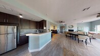 Located at 1175 Pinellas Point Dr S in St. Petersburg, Florida, this single-story home offers a thoughtfully designed open-concept layout that seamlessly connects its primary living spaces. The kitchen is outfitted with rich dark cabinetry, a mosaic tile backsplash, stainless steel appliances, and a curved peninsula with a light countertop — providing both functionality and a striking visual contrast. Wood-look tile flooring flows throughout, tying the interior together with a cohesive, contemporary feel. The living room serves as the heart of the home, anchored by a dramatic floor-to-ceiling white painted brick fireplace with a raised hearth. Soft sage-green walls and a large window framing lush outdoor greenery fill the space with natural light and a calm, coastal ambiance. A ceiling fan adds comfort to the open living area. The dining area is a bright, inviting space with a large picture window overlooking the landscaped yard, ideal for everyday meals or entertaining. A dedicated media room provides a more intimate retreat, featuring recessed lighting, a built-in entertainment center, and rows of leather reclining seating — a true home theater experience. Throughout the home, the consistent use of soft tones, natural light, and thoughtful design details create a relaxed yet polished Florida living environment.