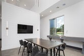 This contemporary single-floor communal space is thoughtfully designed with a blend of modern and transitional elements that create an inviting, upscale atmosphere. Soaring ceilings throughout amplify the sense of openness, while polished concrete floors provide a sleek, cohesive foundation across the entire layout.

A striking focal point is the floor-to-ceiling black and gold marble feature wall, which anchors a built-in wet bar with warm wood cabinetry and dark stone countertops — an elegant and functional centerpiece ideal for entertaining. Adjacent to this, a spacious lounge area is furnished with a plush white sectional sofa, rich cognac leather accent chairs, and a live-edge wood coffee table, all positioned atop a herringbone-patterned area rug.

A white shiplap accent wall with sleek vertical sconce lighting and a wall-mounted flat-screen TV serves as a stylish divider, creating distinct zones within the open floor plan. A dedicated dining and game area features a sturdy high-top table with modern barrel chairs, a mounted television, and a shuffleboard table, all bathed in natural light from large windows overlooking lush greenery.

Recessed lighting throughout and abundant window placement ensure the space is bright and welcoming, with outdoor views adding a refreshing connection to the surrounding landscape.