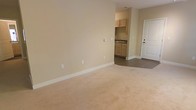 This single-floor space presents a straightforward and functional layout designed for comfortable everyday living. The main living area is finished with plush wall-to-wall carpeting in a warm beige tone, complemented by smooth, neutral-colored walls that create a bright and welcoming atmosphere. Clean white baseboards provide a crisp contrast throughout the space.

Transitioning from the living area, the flooring shifts to a hard surface as the space opens into a compact kitchenette. The kitchenette is outfitted with light wood cabinetry, offering ample storage, and features a countertop with a built-in sink. The cabinetry's natural finish adds a subtle warmth to the functional cooking area.

A paneled entry door with a brushed hardware finish provides access to the unit, contributing to the space's clean and understated aesthetic. The layout flows efficiently between the living and kitchen areas, making practical use of the available footprint. Electrical outlets are conveniently positioned throughout the living area, and a wall-mounted thermostat or control panel is visible, adding to the unit's functional design. Overall, the space is well-maintained, move-in ready, and suited for those seeking a simple, clean, and efficient living environment.