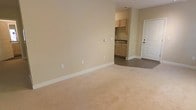 This single-floor space presents a straightforward and functional layout designed for comfortable everyday living. The main living area is finished with plush wall-to-wall carpeting in a warm beige tone, complemented by smooth, neutral-colored walls that create a bright and welcoming atmosphere. Clean white baseboards provide a crisp contrast throughout the space.

Transitioning from the living area, the flooring shifts to a hard surface as the space opens into a compact kitchenette. The kitchenette is outfitted with light wood cabinetry, offering ample storage, and features a countertop with a built-in sink. The cabinetry's natural finish adds a subtle warmth to the functional cooking area.

A paneled entry door with a brushed hardware finish provides access to the unit, contributing to the space's clean and understated aesthetic. The layout flows efficiently between the living and kitchen areas, making practical use of the available footprint. Electrical outlets are conveniently positioned throughout the living area, and a wall-mounted thermostat or control panel is visible, adding to the unit's functional design. Overall, the space is well-maintained, move-in ready, and suited for those seeking a simple, clean, and efficient living environment.