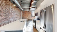 Located on Hertel Avenue in Buffalo, NY, this 2-bedroom, 1-bathroom loft blends industrial heritage with contemporary design. The open-concept living and dining area commands attention with soaring ceilings that expose original heavy timber beams, galvanized ductwork, and corrugated metal deck — a striking nod to the building's industrial past. Polished concrete floors run throughout, reflecting natural light that streams in through large, multi-pane windows framed by original exposed brick walls.

The kitchen is a standout feature, centered around a spacious island with a white quartz countertop and undermount sink. Sleek white cabinetry pairs with stainless steel appliances, including a French door refrigerator, range, and built-in microwave, while a subway tile backsplash adds a clean, modern touch.

The bedrooms offer a calm retreat, with the primary room featuring exposed brick accent walls, generous natural light, and a stylish sliding glass-panel barn door. The bathroom is finished with white subway tile in the walk-in shower, a modern floating vanity with an illuminated mirror, and polished concrete flooring. In-unit laundry adds everyday convenience, completing this well-appointed urban loft.