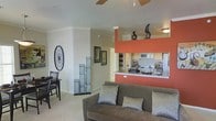 This single-story residence features an open-concept layout that integrates the living, dining, and kitchen areas into one cohesive space. The living area is anchored by a plush taupe sofa adorned with patterned throw pillows, set atop wall-to-wall carpeting that adds warmth underfoot. A ceiling fan overhead provides both function and a finishing touch to the comfortable seating area.

The kitchen is a standout feature, distinguished by a bold coral-orange accent wall and raised breakfast bar with a light stone-toned countertop. White cabinetry and subway tile backsplash complement the stainless steel appliances, including a microwave and refrigerator. Decorative sculptures displayed above the upper cabinetry add an artistic element to the space.

Adjacent to the living area, a small dining nook accommodates a dark wood table and chairs, while decorative wall art and sculptural accents throughout lend personality to the neutral, beige-toned walls. Crown molding adds a refined architectural detail. The overall ambiance is warm and inviting, with a balanced mix of bold color and neutral tones creating a visually dynamic yet comfortable environment.
