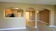 This two-floor home showcases a thoughtfully designed interior that balances openness with architectural character. The main living area is defined by a series of graceful arched doorways and passageways that create a sense of flow between rooms while adding a refined architectural detail to the space. Warm, neutral-toned walls complement the light-colored carpet underfoot, contributing to a cohesive and inviting atmosphere throughout.

The kitchen is accessible via an arched opening and features a raised breakfast bar that separates it from the living area while maintaining visual connectivity. Appliances including a refrigerator and microwave are visible, and cabinetry lines the walls to provide ample storage. An additional arched passage leads toward what appears to be an entryway, where a front door with decorative glass panels allows natural light to filter into the interior.

A secondary living or family area is accessible through another arch and is equipped with a ceiling fan, enhancing comfort and air circulation. White baseboards provide clean contrast against the neutral walls, and recessed lighting supplements the overall illumination. The layout encourages an easy, connected movement between the home's key areas.