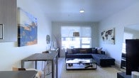 This single-floor space offers a sleek, modern aesthetic defined by clean lines, crisp white walls, and an open-concept layout that maximizes the sense of space. The living area is the centerpiece of the unit, anchored by a plush sectional sofa in cool grey tones, accented with warm-hued throw pillows that add a welcoming contrast. A glass-topped coffee table sits atop a soft, textured area rug, completing the comfortable yet refined seating arrangement. A large window with roller shades floods the room with natural light, creating a bright and airy atmosphere throughout the day. A statement arc floor lamp adds a sculptural design element while providing warm task lighting in the evenings. Artwork adorns the walls, including a bold abstract blue-and-gold canvas on one side and a world map print on the other, lending the space a well-traveled, culturally inspired character. A bar-height dining table with industrial-style seating provides a distinct dining or workspace area, seamlessly integrated into the open floor plan. The overall ambiance is contemporary and well-curated, blending functional living with thoughtful design details that make this space both practical and visually engaging.