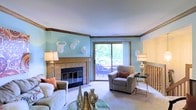 This two-floor space offers a thoughtfully designed interior with a living room that serves as its centerpiece. The room is finished with soft blue walls accented by warm wood crown molding that runs along the upper perimeter, lending a classic and polished character to the space. A tile-surround fireplace with a light wood mantle anchors one wall, providing both a functional and decorative focal point. Neutral carpet flooring extends throughout, contributing to the overall warmth and comfort of the room. Natural light floods the space through a large sliding glass door, which also provides direct access to an outdoor balcony or deck where seating is visible, surrounded by mature trees. The indoor-outdoor connection enhances the sense of openness. Decorative white wall-mounted sculptures add a distinctive, eclectic touch above the fireplace. The living room comfortably accommodates seating arrangements suited for relaxing or entertaining, with a glass-top coffee table completing the setup. The space spans two floors, suggesting additional rooms with varied functions that complement this bright, characterful main living area.