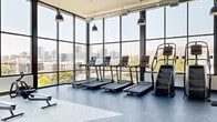 This modern fitness center occupies a single, expansive floor and is designed to deliver a premium workout experience. The most striking architectural feature is the continuous floor-to-ceiling curtain wall of windows that lines multiple sides of the space, flooding the interior with abundant natural light and framing panoramic views of the surrounding cityscape and hillside beyond. Exposed ductwork runs along the white ceiling, complemented by industrial-style pendant lighting that adds a contemporary, urban aesthetic.

The flooring is a striking combination of white marble-look and dark slate-toned tile, creating distinct visual zones throughout the space. A textured brick accent wall anchors the interior side of the gym, adding warmth and character alongside the sleek modern finishes.

The fitness floor is comprehensively equipped, featuring multiple treadmills, stair climbers, stationary and spin bikes, rowing machines, and a full Matrix cable and weight machine setup. A dedicated free weights area with racks, dumbbells, and adjustable benches is positioned near full-length mirrors. The layout provides ample open floor space between equipment zones, supporting a variety of workout styles. A glass-paneled entrance with keycard access completes the professional, polished design of this well-appointed facility.