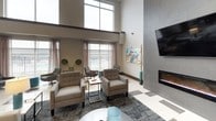 This well-appointed single-floor amenity space makes a striking first impression with its expansive lobby, showcasing a bold geometric black-and-white tile floor that extends throughout much of the space. Soaring double-height ceilings in the main entry and lounge areas lend an open, airy quality, complemented by a mezzanine-level overlook with sleek metal railings and a grand staircase anchoring the central corridor.

The lounge area is a standout feature, designed for comfort and socializing. Plush upholstered armchairs and accent seating are arranged around a modern linear fireplace set into a floor-to-ceiling tiled feature wall. A wall-mounted flat-screen television adds a media-friendly element to the space. Floor-to-ceiling windows draped with sheer curtains flood the room with natural light and offer direct views of the outdoor pool area, creating a seamless indoor-outdoor connection.

A secondary seating nook near the windows features eclectic patterned accent chairs and teal ottomans, providing a casual and colorful counterpoint to the more neutral lounge furnishings.

The fully equipped kitchen is finished with dark espresso cabinetry, quartz countertops, stainless steel appliances including a range and refrigerator, a geometric hexagonal tile backsplash, and a deep undermount sink — combining functionality with refined aesthetics throughout.