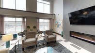 This well-appointed single-floor amenity space makes a striking first impression with its expansive lobby, showcasing a bold geometric black-and-white tile floor that extends throughout much of the space. Soaring double-height ceilings in the main entry and lounge areas lend an open, airy quality, complemented by a mezzanine-level overlook with sleek metal railings and a grand staircase anchoring the central corridor.

The lounge area is a standout feature, designed for comfort and socializing. Plush upholstered armchairs and accent seating are arranged around a modern linear fireplace set into a floor-to-ceiling tiled feature wall. A wall-mounted flat-screen television adds a media-friendly element to the space. Floor-to-ceiling windows draped with sheer curtains flood the room with natural light and offer direct views of the outdoor pool area, creating a seamless indoor-outdoor connection.

A secondary seating nook near the windows features eclectic patterned accent chairs and teal ottomans, providing a casual and colorful counterpoint to the more neutral lounge furnishings.

The fully equipped kitchen is finished with dark espresso cabinetry, quartz countertops, stainless steel appliances including a range and refrigerator, a geometric hexagonal tile backsplash, and a deep undermount sink — combining functionality with refined aesthetics throughout.