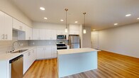 This thoughtfully designed residence offers three bedrooms, two bathrooms, and a functional single-floor layout that maximizes every square foot. The open-concept living and kitchen area serves as the heart of the home, featuring warm hardwood-style flooring, recessed lighting, and large windows that invite abundant natural light. The kitchen is appointed with crisp white cabinetry, a subway tile backsplash, stainless steel appliances — including a refrigerator, range, microwave, and dishwasher — and a central island with pendant lighting, providing both prep space and a casual dining option. Built-in open shelving adds practical storage near the main living area.

The bedrooms are carpeted for comfort, each featuring recessed lighting and windows with roller shades offering outdoor views of the surrounding community. The primary bedroom includes a ceiling fan and a dedicated closet with built-in shelving. The bathrooms are finished with clean white vanities, modern fixtures, and large mirrors. A dedicated laundry room houses a full-size front-loading washer and dryer set with overhead wire shelving. Two hallways connect the space efficiently, and a separate closet provides additional storage. The overall aesthetic is clean, contemporary, and move-in ready.