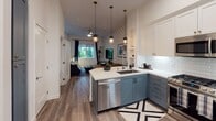 Located at 1961 E Dyer Rd in Santa Ana, California, this single-floor unit offers a well-appointed, open-concept layout that seamlessly connects the kitchen, dining, and living areas. The kitchen is a standout feature, showcasing two-tone cabinetry with slate-blue lower cabinets and crisp white uppers, complemented by white subway tile backsplash, quartz countertops, stainless steel appliances, and a peninsula with bar seating. Stylish pendant lighting with warm brass accents hangs above, adding a contemporary touch. The dining area is anchored by a rustic wood table set against a bold geometric-patterned accent wall, creating a striking visual focal point. The living room is bright and welcoming, with hardwood-style flooring, a ceiling fan, and a large window framed by dark curtains that invites natural light and views of lush tropical greenery outdoors. The bathroom is clean and modern, featuring slate-blue vanity cabinetry matching the kitchen, quartz countertops, a large frameless mirror, and a tiled shower-tub combination. Throughout the space, consistent design choices — including coordinated cabinetry finishes, warm flooring, and curated décor — create a cohesive, contemporary aesthetic that is both functional and visually appealing.