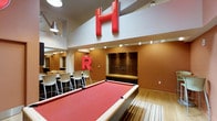 This single-floor recreational lounge is designed to serve as a lively social gathering space with a strong, energetic aesthetic. The centerpiece of the room is a full-size pool table with a striking red felt surface, anchoring the space and inviting active engagement. Soaring double-height ceilings create an open, airy atmosphere, while oversized bold red letter installations mounted on the upper walls add a distinctive branded character to the interior. Warm terracotta-toned walls contrast with crisp white ceiling elements, lending the space a modern yet welcoming ambiance. A bar-height seating area lines one side of the room, featuring sleek white bar stools and a dark countertop — ideal for casual conversation and socializing. Adjacent to the pool table, a built-in wooden cabinetry unit with recessed display shelving serves as a stylish focal point along the back wall. Light hardwood flooring runs throughout, complemented by a patterned area rug beneath the pool table that adds texture and visual interest. Pendant lighting and recessed fixtures provide well-distributed illumination throughout the space, enhancing its inviting, social character. Overall, the lounge strikes a balance between playful design and functional entertainment amenities.