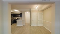 This single-floor residence offers a clean, open-concept layout that seamlessly connects the living area to the kitchen, creating a spacious and functional environment. Light-toned hardwood flooring runs throughout, lending warmth and continuity to the space.

The kitchen is a standout feature, showcasing dark cabinetry that contrasts boldly against the light surroundings. A mosaic tile backsplash adds a decorative touch, while stainless steel appliances — including a microwave and range — provide a modern, polished look. A raised peninsula counter serves as both a workspace and an informal dining or gathering spot, opening directly to the main living area.

An elegant arched doorway frames the transition between the entryway corridor and the main living space, adding an architectural detail that softens the overall design. The entryway area includes a panel door leading to the exterior, flanked by additional interior doors — one featuring classic louvered panels, likely a utility or storage closet.

Track lighting installed along the ceiling of the living area provides focused, adjustable illumination, complementing the flush-mount fixtures found elsewhere. Neutral beige walls and white trim throughout create a bright, inviting ambiance. Crown molding adds a refined finishing touch, elevating the overall character of the interior.