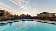 Located at 14158 S Hilltop Ln in Lockport, Illinois, this outdoor pool area serves as a central amenity space within a residential community. The generously sized swimming pool features classic geometric design with multiple entry points via stainless steel ladder rails and a wide step entry, making it accessible from several sides. The pool's clear, blue-tinted water reflects the open sky, creating a bright and inviting atmosphere throughout the day and into the evening hours.

Surrounding the pool is a broad concrete sundeck that provides ample room for lounging and socializing. Numerous chaise lounge chairs are arranged along the perimeter, offering comfortable spots for relaxation in the sun. Tall black lamp posts are positioned around the deck, providing lighting for extended evening use, while a wrought iron perimeter fence encloses the area for safety and security.

Adjacent to the pool, a well-appointed single-story clubhouse building constructed with brick and stone accents serves as a community hub. Large windows and glass doors fill the structure with natural light, and an outdoor patio area in front features bistro-style tables and chairs with market umbrellas, ideal for casual outdoor dining or gatherings. Together, these elements create a cohesive, resort-style outdoor environment backed by sweeping views of the surrounding residential buildings and open sky.