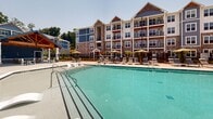 Located at 640 Lakeside Dr in Canton, GA, this thoughtfully designed outdoor amenity space offers a resort-style experience for its community. At its centerpiece is a generously sized swimming pool with crystal-clear water, flanked by a broad concrete sundeck furnished with lounge chairs, including in-pool tanning ledge loungers perfect for relaxing in the Georgia sun. The pool area is enclosed by elegant black iron fencing, complemented by manicured landscaping featuring evergreen shrubs and mulched garden beds that add a polished, welcoming atmosphere.

Adjacent to the pool, a vibrant red pergola anchors a dedicated outdoor entertaining zone, complete with an outdoor kitchen and bar seating, ideal for gatherings. Nearby, a covered lounge nook framed by warm cedar-clad walls provides an intimate, semi-private seating area furnished with plush sofas and accent chairs in neutral and teal tones. Additional patio seating with dining tables and chairs is distributed throughout the space, offering multiple areas for socializing or relaxing.

The backdrop is defined by the community's multi-story residential buildings, showcasing a charming mix of craftsman-style architectural elements including stone accents, blue siding, gabled rooflines, and private balconies. The overall ambiance is bright, open, and resort-inspired.