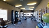 This fitness center is designed to support a well-rounded workout routine within a single-floor layout. The room is finished with neutral gray walls and durable dark flooring, creating a functional and focused environment. Overhead, a series of rectangular fluorescent fixtures provide consistent, bright illumination throughout the space, complementing the natural light that filters in through a row of windows along the far wall. These windows not only brighten the room but also create an open, airy atmosphere conducive to exercise.

The facility is outfitted with a selection of cardio equipment, including treadmills, an elliptical trainer, and a stationary recumbent bike, all positioned to take advantage of the natural light from the windows. A multi-function cable weight machine and an adjustable weight bench are situated on the left side of the room, offering strength training options. Two wall-mounted flat-screen televisions are positioned at opposite ends of the room, providing entertainment during workouts. The clean white baseboards and organized layout contribute to a tidy, well-maintained atmosphere that supports an effective fitness experience.