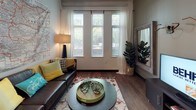 This single-floor unit is designed to accommodate multiple residents, offering a thoughtful layout that balances private and shared living spaces. The open-concept dollhouse view reveals a well-organized floor plan with distinct zones for sleeping, studying, cooking, and relaxing.

Each bedroom is compact yet efficiently arranged, featuring a platform bed with integrated storage drawers beneath, a dedicated desk with an ergonomic chair, and a window with blinds and curtains that allow natural light while maintaining privacy. Carpet tile flooring adds warmth underfoot, and wall art personalizes each room.

The bathrooms feature dark concrete-style flooring, wall-mounted storage cabinets, and clean white fixtures, maintaining a modern, minimalist aesthetic throughout.

The shared living area is anchored by tall windows that flood the space with natural light and offer views of surrounding greenery. Wood-style flooring, a leather sofa, and a round coffee table create an inviting communal atmosphere.

The kitchen is sleekly designed with dark lower cabinetry, wood-toned upper cabinets, light countertops, and a full suite of stainless steel appliances, including a gas range, dishwasher, and microwave, providing a fully functional cooking space.