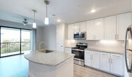Located in Fort Myers, FL, this one-bedroom, one-bathroom residence offers a thoughtfully designed open-concept layout. The combined kitchen, dining, and living area serves as the heart of the home, featuring light wood-style flooring and a seamless flow enhanced by natural light from a sliding glass door that opens onto a private patio. The kitchen is a standout feature, equipped with white shaker-style cabinetry extending to the ceiling, granite countertops, a curved island with an undermount sink, stainless steel appliances including a refrigerator, electric range, and built-in microwave, plus a classic subway tile backsplash. Pendant lighting over the island adds a modern touch. The living area includes a ceiling fan and direct patio access with outdoor views. The bedroom is a comfortable retreat with plush carpeting, a large window overlooking the surrounding landscape, and generous closet space — the unit includes five closets in total. The bathroom mirrors the home's clean aesthetic with a white vanity, granite countertop, and wood-style flooring. A hallway connects the main areas efficiently. The overall ambiance is bright, clean, and contemporary, reflecting a modern residential design suited to the Fort Myers lifestyle.