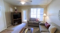 This single-floor space offers a welcoming and functional layout designed for comfortable everyday living. The living room serves as the heart of the home, featuring plush wall-to-wall carpeting that adds warmth underfoot. A ceiling fan with an integrated light fixture sits centrally overhead, providing both air circulation and ambient lighting throughout the space.

Natural light floods the room through a large window fitted with horizontal blinds, allowing for adjustable light control while offering a view of the outdoors. A glass-paneled door provides direct access to what appears to be a balcony or patio, further enhancing the sense of openness and connection to the exterior.

The living area is thoughtfully arranged with comfortable seating, including a loveseat and sofa accented with decorative pillows, centered around a rustic coffee table. A wooden media console with open shelving provides practical storage and display space alongside the entertainment setup. The neutral wall tones and warm carpeting create a cozy, inviting ambiance that makes the space feel both relaxed and functional. Overall, the layout is efficient and well-suited for everyday living and entertaining.