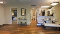 This single-floor residence offers a cohesive and welcoming layout defined by rich hardwood flooring that flows seamlessly throughout the living areas. The open floor plan connects the dining area, hallway, and adjacent rooms, creating a sense of spaciousness and ease of movement. The dining area is furnished with a glass-top table and upholstered seating, set against a backdrop of warm, neutral-toned walls that contribute to the overall inviting ambiance. A decorative console table with a mirrored frame and curated coastal accents adds a stylish focal point along the hallway, complemented by a wall-mounted mirror that enhances depth and light. The bedroom, visible through an open doorway, is generously proportioned and features soft, neutral bedding. A full bathroom down the hall is bright and clean, with classic fixtures and a tidy, functional layout. The kitchen is partially visible and includes stainless steel appliances and stone countertops, suggesting a well-equipped cooking space. Recessed and flush-mount lighting throughout ensures the interiors remain bright and functional. The neutral color palette, warm wood tones, and coastal-inspired décor elements combine to create a refined yet comfortable living environment.