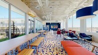 This dynamic single-floor amenity space is designed to inspire social interaction and leisure through bold, contemporary design. The open layout is anchored by a striking life-size chess set displayed on a black-and-white checkered floor inlay, serving as both a functional game area and a dramatic visual centerpiece. Throughout the space, a vibrant terrazzo-style patterned carpet in blue, white, and earth tones ties together the various seating zones. Long communal wood tables with bench seating invite casual gathering, while plush lounge areas feature an eclectic mix of blue, red, and neutral upholstered chairs and sofas arranged around low coffee tables. A cozy reading nook offers a pair of cream wingback chairs accented with bold red cushions, positioned beside a floor-to-ceiling window framing open sky views. Distinctive pendant lighting — including large rattan globe fixtures and oversized blue drum shades — adds warmth and character overhead, while exposed structural ceilings with white-painted ductwork lend an industrial-modern aesthetic. Floor-to-ceiling glass walls and oversized windows flood the interior with abundant natural light and provide sweeping views of the surrounding outdoor terrace and community. Colorful geometric murals and curated artwork on deep navy accent walls complete the vibrant, contemporary atmosphere.