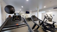 This fitness center offers a comprehensive workout environment designed to support a wide range of exercise routines. The space is finished with clean white walls and a neutral carpet floor, creating a bright and motivating atmosphere enhanced by recessed lighting throughout the ceiling. Multiple treadmills with digital display consoles are prominently featured, providing ample cardio options for users. A dedicated free weights area lines one wall, with a full rack of dumbbells organized for easy access alongside a multi-functional cable and weight machine, catering to strength training needs. A wall-mounted flat-screen television is positioned to keep users entertained during workouts. Exercise and stability balls are also available, rounding out the functional fitness offerings. A large mirror on one wall expands the sense of space and allows users to monitor their form. The layout is thoughtfully arranged to maximize usable floor area while maintaining clear pathways between equipment. Additional doors suggest access to changing or utility areas, adding to the overall convenience of the facility. The space is well-maintained and efficiently organized, making it a practical and fully functional fitness resource.