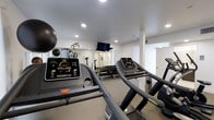 This fitness center offers a comprehensive workout environment designed to support a wide range of exercise routines. The space is finished with clean white walls and a neutral carpet floor, creating a bright and motivating atmosphere enhanced by recessed lighting throughout the ceiling. Multiple treadmills with digital display consoles are prominently featured, providing ample cardio options for users. A dedicated free weights area lines one wall, with a full rack of dumbbells organized for easy access alongside a multi-functional cable and weight machine, catering to strength training needs. A wall-mounted flat-screen television is positioned to keep users entertained during workouts. Exercise and stability balls are also available, rounding out the functional fitness offerings. A large mirror on one wall expands the sense of space and allows users to monitor their form. The layout is thoughtfully arranged to maximize usable floor area while maintaining clear pathways between equipment. Additional doors suggest access to changing or utility areas, adding to the overall convenience of the facility. The space is well-maintained and efficiently organized, making it a practical and fully functional fitness resource.